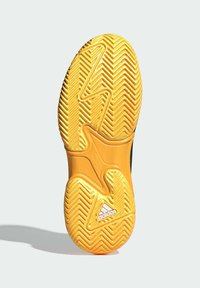 Žlutá podrážka bot Adidas se zigzagovým vzorem a bílým logem Adidas poblíž paty, vrhající stín na neutrálním pozadí.