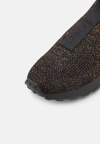 Gros plan sur l'avant d'une basket en maille noire et bronze, avec une semelle texturée et une étiquette noire de marque sur le tissu en mesh supérieur.