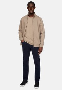 Chaqueta beige con cremallera sobre una camiseta beige, combinada con pantalones azul marino oscuros y zapatillas negras, con una textura suave y un diseño minimalista.