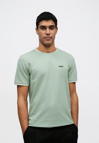 T-shirt de manga curta verde claro com gola redonda, apresentando detalhes listrados contrastantes na gola e nas mangas, e um pequeno logótipo preto.