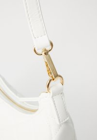 Weißer gesteppter Lederhandtaschenriemen mit goldener Hardware und einem Verschluss. Die Textur zeigt glattes Leder; die Nähte sind am Riemen sichtbar.