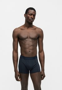 Άνδρας μοντέλο στέκεται μπροστά σε απλό φόντο φορώντας σκούρα εφαρμοστά μπόξερ, με ουδέτερο έκφραση προσώπου και χαλαρή στάση.