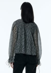 Zwart-wit gebloemde blouse met doorschijnende lange mouwen, geplooide details bij de halslijn en manchetten, gecombineerd met donkere jeans.