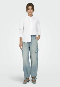 Camicia bianca con bottoni e maniche lunghe, abbinata a pantaloni cargo blu chiaro. I pantaloni presentano tasche laterali e caviglie arricciate. Tacchi verdi.