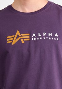 Fialové tričko s krátkým rukávem vyrobené z bavlny, s velkým oranžovým logem obsahujícím abstraktní "A" a křídly vedle textu "ALPHA INDUSTRIES."