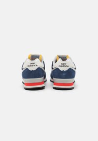 Baskets de sport en velours bleu avec des accents blancs et une semelle intermédiaire rouge. Présentent un talon rembourré et un détail jaune au niveau du col. Marque "New Balance" visible.