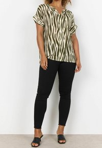 Top a maniche corte con scollo a V caratterizzato da un motivo astratto verde e crema, abbinato a jeans skinny neri e sandali neri open-toe.