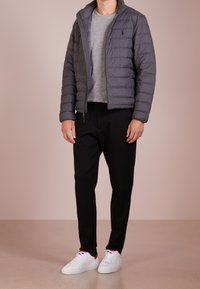 Grå pufferjacka med dragkedja, strukturerat tyg och en subtil logotyp. Matchas med en grå skjorta, svarta byxor och vita sneakers.