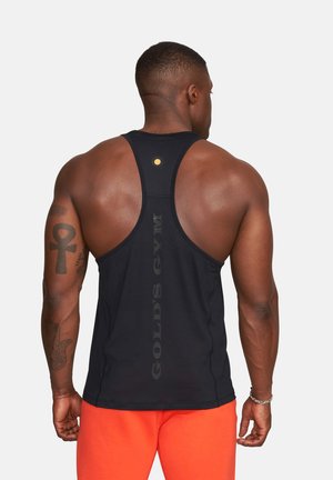 Muskularny mężczyzna z tatuażami, ubrany w czarny tank top Gold's Gym oraz jasnopomarańczowe szorty, stoi plecami do widza na jednolitym tle.