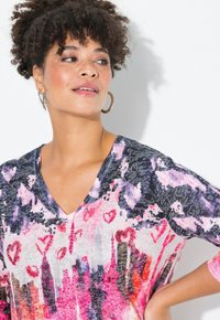 Flerfärgad topp med V-ringning, korta ärmar och en blommig och abstrakt design i rosa, lila och svart, med dekorativa detaljer.