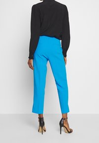 Osoba w jasnoniebieskich, krótkich spodniach, czarnej bluzce z długim rękawem oraz czarnych sandałach na wysokim obcasie, stojąca na jednolitym tle.