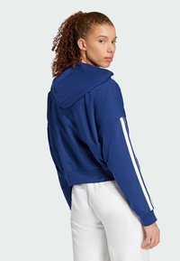 Skraćena plava dukserica s velikom kapuljačom, bijelim bočnim trakama i rebrastim manžetama, u kombinaciji s bijelim hlačama. Glatka tekstura tkanine.