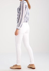 Camisa de botones a rayas azul y blanca, jeans slim-fit blancos y slippers marrón claro. La camisa tiene mangas largas y puños con detalles de botones.