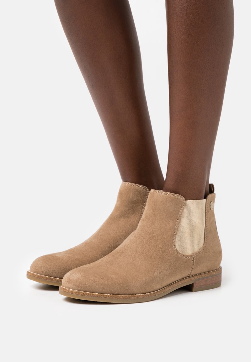 Bottines en daim beige avec panneaux latéraux élastiques et talons bas en bois, portées sur des jambes à la peau foncée sur fond blanc.