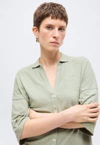 Άτομο με κοντά καστανά μαλλιά, φοράει ανοιχτοπράσινο πουκάμισο με κουμπιά και χρυσά σκουλαρίκια, με σταυρωμένα τα χέρια, κοιτάει μπροστά με ουδέτερη έκφραση.