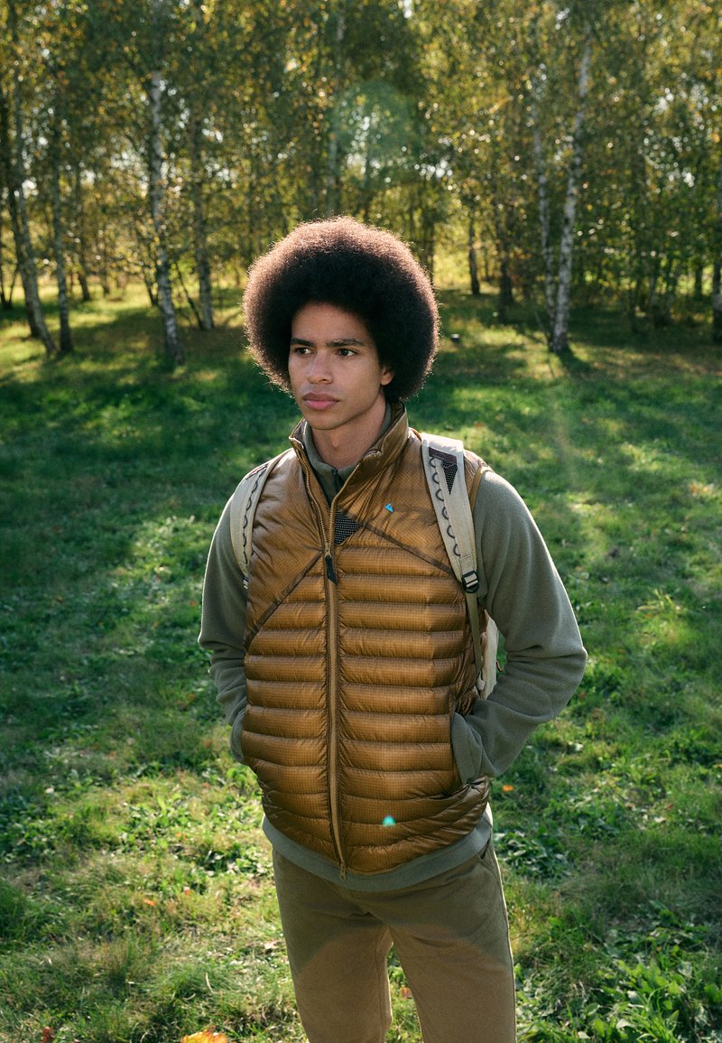 Brown puffy vest with horizontal stitching, worn over a gray long-sleeve shirt, paired with light-colored pants. Outdoor setting with greenery.