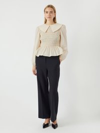 Helles beige Peplum-Top mit großem Kragen, gerafftem Oberteil und langen Puffärmeln, kombiniert mit schwarzen maßgeschneiderten Hosen und spitzen schwarzen Schuhen.
