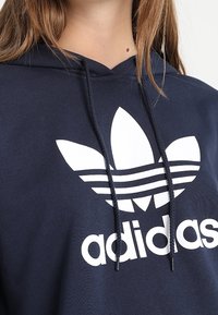 Mörkblå hoodies i bomull med en stor vit Adidas-logotyp med tre ränder och en blomliknande design på bröstet.