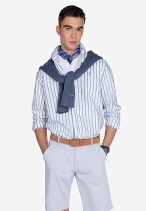 Camisa de botones a rayas azul claro y blanca con un suéter de punto azul marino drapeado alrededor del cuello, combinado con pantalones cortos claros y un cinturón marrón.