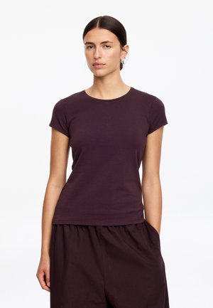 Femme aux cheveux foncés en chignon bas portant des boucles d’oreilles créoles argentées, un t-shirt ajusté violet foncé à manches courtes et un pantalon ample assorti.