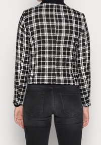 Musta-valkoinen ruudullinen lyhyt takki pitkillä hihoilla, teksturoidusta kankaasta, pyöreällä kauluksella ja yksinkertaisilla ompelusyillä. Päällä mustat farkut.