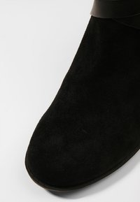 Musta mokkanahkainen nilkkuri pyöreällä kärjellä, jossa yläosassa musta nahkahihna ja litteä, kuvioitu pohja.