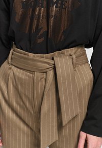 Beige streepjasje met een brede zelfbindriem, gedragen met een zwart overhemd met lange mouwen en een reflecterend abstract patroon.