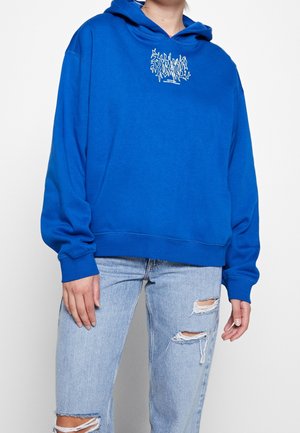 Person wearing a bright blue hoodie with white text on chest and light blue ripped jeans, standing against a plain white background.