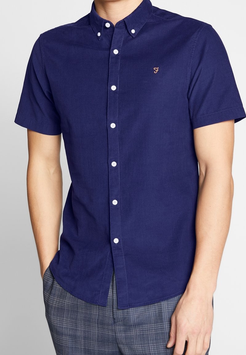 Homme portant une chemise bleu marine à manches courtes boutonnée avec un petit logo brodé sur la poitrine et un pantalon gris à carreaux.