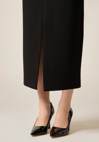 Falda lápiz negra con una abertura frontal, combinada con tacones altos de charol negro con punta afilada y acabado liso.