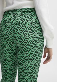 Grüne gemusterte Hosen mit schwarzen geometrischen Formen, elastischem Bund, seitlichen Taschen und einer glatten Stofftextur.