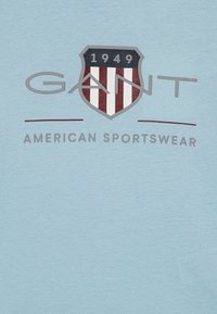 Una camiseta de color azul claro presenta un logo de escudo con franjas rojas y blancas, texto gris "GANT" y "AMERICAN SPORTSWEAR," y el año "1949."