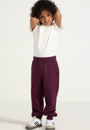 Barn med krøllet hår iført en hvid t-shirt, lilla sweatpants og hvide sneakers, stående med hænderne bag hovedet mod en ensfarvet baggrund.