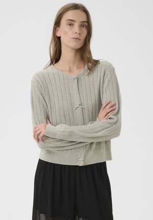 Ženska z dolgimi lasmi, oblečena v svetlo sivi pulover iz rebrastega džersija in črno krilo, z rokami prekrižanimi, nevtralen izraz, enobarvno svetlo ozadje.