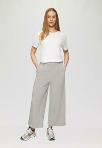 Helle graue, weite Hose mit zwei vorderen Taschen, kombiniert mit einem schlichten weißen T-Shirt und silbernen Turnschuhen, vor einem neutralen Hintergrund.