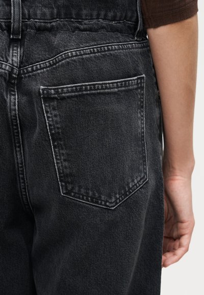 Κοντινό πλάνο ενός ατόμου που φοράει μαύρα τζιν παντελόνια, δείχνοντας την πίσω τσέπη και μέρος ενός καφέ μανικιού στο δεξί χέρι.