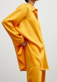 Femme portant une chemise ample à manches longues en satin orange vif avec un pantalon large assorti, main glissée dans la poche, sur un fond uni.