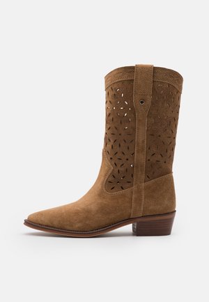 Botín de ante marrón con diseños de corte floral, puntera afilada, tacón de madera y detalles de costura en la parte superior y los laterales.