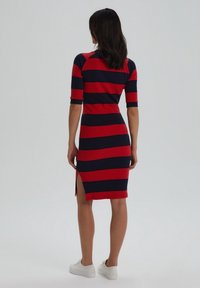 Donna in piedi, che indossa un vestito a righe orizzontali rosso e nero fino al ginocchio con maniche corte e sneaker bianche, di spalle rispetto alla fotocamera.