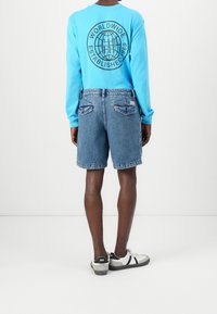 Camisa azul de manga larga con un gráfico circular negro en la parte posterior, combinada con pantalones cortos de mezclilla azul y zapatillas blancas con detalles negros.