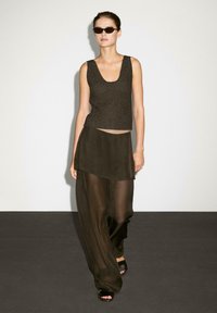 Modelka kráčí v elegantním tmavém outfitu, který zahrnuje přiléhavý top a průsvitné, splývavé kalhoty, doplněné stylovými slunečními brýlemi na minimalistickém pozadí.