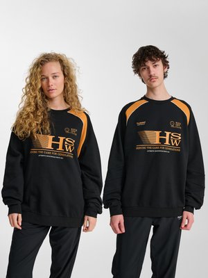 To unge voksne iført matchende sorte og orange sportssweatshirts med teksten "HSW" og "Kick Off Club", står foran en ensfarvet baggrund.