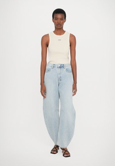 Lichtbeige mouwloos topje met logo, gecombineerd met lichtblauwe, oversized jeans met een hoge taille en subtiele naden aan de voorkant. Zwarte sandalen met bandjes.