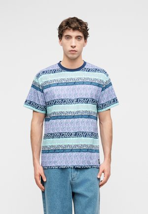 Bărbat tânăr purtând un tricou cu mânecă scurtă, cu model în tonuri de albastru și violet, asortat cu blugi bleu, stând în picioare pe un fundal simplu.