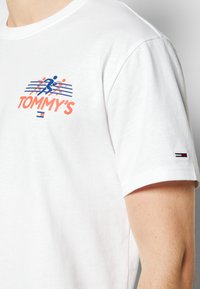 Hvid bomulds t-shirt med et farverigt grafisk design, der har "TOMMY'S" tekst, blå og orange accenter samt et lille logo på ærmet.