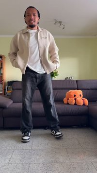 Beige knapjakke over en hvid t-shirt, sorte jeans og sorte sneakers. En orange plysjblekksprut sidder på en mørk sofa i baggrunden.