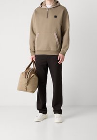 Beżowa bluza z kapturem na suwak, z kieszenią kangurką i kontrastującymi akcentami materiałowymi. Czarne spodnie i beżowa torba typu duffle z brązowymi uchwytami. Białe trampki.