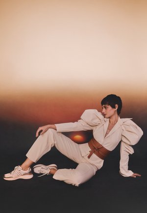 Femme allongée sur le sol portant un tailleur crème à manches bouffantes, une large ceinture marron et des baskets, sur un fond dégradé marron et noir.