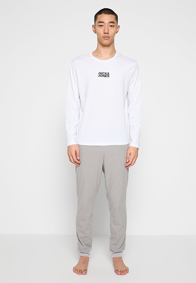T-shirt bianca a maniche lunghe con un logo nero sul petto, abbinata a pantaloni grigi chiari con polsini elastici alle caviglie.