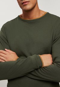 Ciemnozielony sweter z okrągłym dekoltem, z jasnobrązowymi żebrowanymi akcentami przy dekolcie i mankietach. Gładki, teksturowany materiał.